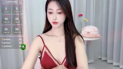 Media: Video of an East Asian woman with long black hair in a red lace camisole, wearing a gold necklace. She stands in a minimalist room with white curtains and a pink stuffed pig.