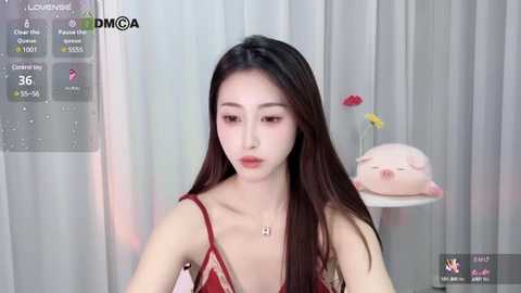 Media: A video of a young East Asian woman with long black hair and fair skin, wearing a red lace camisole. She's seated indoors, surrounded by soft lighting and a pink stuffed animal.