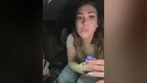 Media: Video of a young woman with light skin, brown hair, and a neutral expression, holding a yellow and purple vibrator, sitting in a dimly lit car.