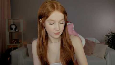 Video of a young, fair-skinned redhead woman with long, straight hair, wearing a sleeveless top, sitting on a white couch in a cozy, dimly lit living room.