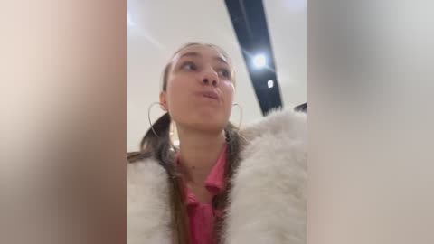 Media: Video of a young woman with light skin, pigtails, and a pink blouse, wearing a white faux fur coat, blowing a kiss in a dimly lit room.