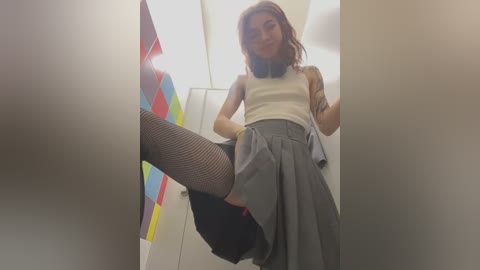 Media: Video of a slender, fair-skinned woman with shoulder-length brown hair in a white sleeveless top and gray pleated skirt, wearing fishnet stockings and black heels, posing in a brightly lit restroom with colorful abstract art in the background.