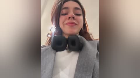 Media: Video of a smiling young woman with medium skin tone and straight, shoulder-length brown hair, wearing a grey blazer over a white top, with large black over-ear headphones around her neck.