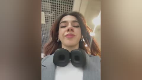 Media: Video of a young woman with light skin, dark hair, and closed eyes, wearing a gray jacket and black headphones. Background shows a grid pattern and a blurry light bulb.