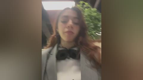 Video of a young woman with long, reddish-brown hair, wearing a white shirt and a gray blazer with black bow tie, standing indoors with a potted plant and fluorescent lighting in the background.