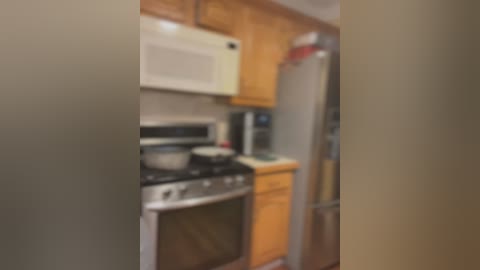 Media: Blurred video of a small, modern kitchen with stainless steel appliances, wooden cabinets, a microwave, and a refrigerator, viewed from a slightly off-center angle.