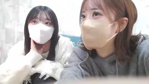 Media: Video of two East Asian women in surgical masks, one with long black hair, the other with light brown hair, both wearing white coats. Background shows a blurred, white and gray room.