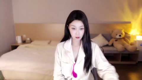 Media: Video of a young Asian woman with long black hair, wearing a white robe with a pink tie, standing in a minimalist bedroom with a beige headboard, teddy bear, and warm lighting.