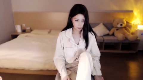 Media: A video of an Asian woman with long black hair, fair skin, wearing a white hoodie, sitting on a bed in a modern, cozy bedroom with beige walls, a teddy bear, and a lit lamp.