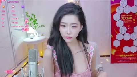 Media: A video of an East Asian woman with long black hair, wearing a pink floral dress, sitting in a brightly lit room with a white sink, pink flowers, and a red poster with Chinese text.