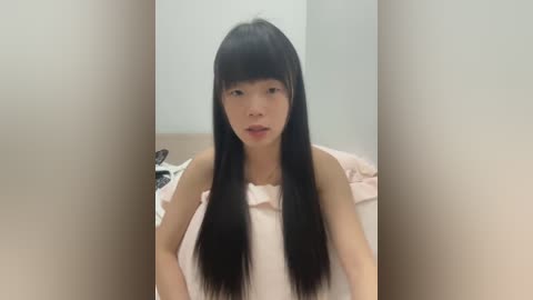 Media: A video of an Asian woman with long black hair and bangs, wearing a light pink off-shoulder top, sitting on a bed with white sheets in a minimalistic room.