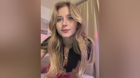 Video of a young Caucasian woman with long, wavy blonde hair, fair skin, and a slender build, wearing a black top, posing indoors with beige curtains and pink bedding in the background.