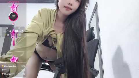 Media: A video of an East Asian woman with long black hair, wearing a green blouse and revealing a black bra, leaning forward, taken indoors with a video camera.