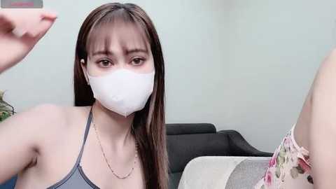 Media: A video of a young East Asian woman with long brown hair, wearing a grey tank top and white face mask, sitting on a grey couch, holding a phone to take a selfie.