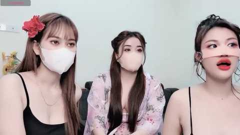 Media: Three young East Asian women, masked and wearing floral-patterned robes, sit closely together on a couch in a modern living room.