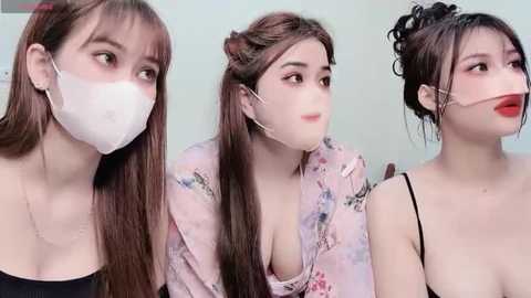 Media: Video of three young Asian women with fair skin, wearing face masks, floral tops, and black spaghetti straps, against a pale green background.