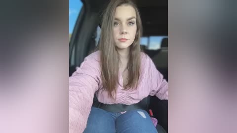 Media: Video of a young woman with long brown hair, wearing a pink ribbed sweater and blue jeans, seated in a car with a blurred background.