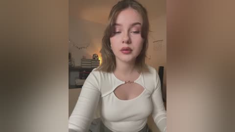 Media: Video of a young Caucasian woman with fair skin, shoulder-length light brown hair, wearing a white top with a keyhole design. She has a neutral expression. Background includes a cozy room with a string of lights and a wall-mounted clock.