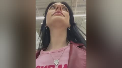 Media: Video of a woman with long black hair, wearing glasses, a pink t-shirt, and a necklace, looking up with a slight smile. Background shows an office with white ceiling tiles and fluorescent lights.