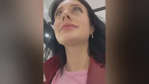 Media: Video of a fair-skinned woman with long black hair, wearing a pink shirt and red jacket, with a small facial piercing, looking upward in an indoor setting.