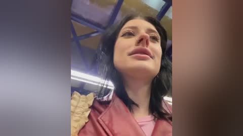 Media: Video of a woman with straight black hair, light skin, and a slight smile, wearing a red jacket and a pink shirt, taken from an upward angle in a subway car with blue and yellow tiles.