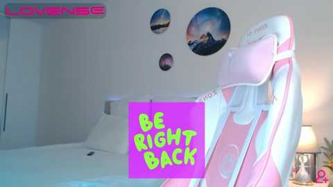 Video of a minimalist bedroom with a white bed, pink and white polka-dotted gaming chair, and \"BE RIGHT BACK\" neon text.