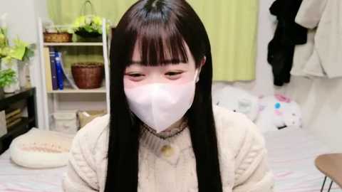 Media: Video of an East Asian woman with long black hair, wearing a white mask, beige sweater, and a necklace, in a cozy bedroom with green curtains, a shelf, and a plush toy.