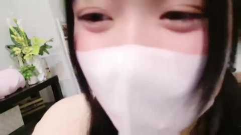 Media: Video of a young Asian woman with long black hair, wearing a white face mask, sitting indoors. Background includes a vase with flowers and a black table.