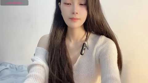 Media: Video of a young East Asian woman with long, straight, dark brown hair, wearing a white off-shoulder knitted sweater, looking serene with closed eyes, set against a plain, light-colored background.