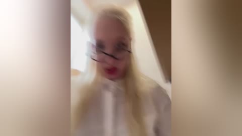 A blurred video of a blonde woman with red lipstick, wearing glasses, and a white shirt, possibly in a bathroom with a white towel in the foreground.