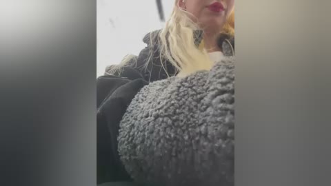 Media: Video of a blonde woman with red lipstick, wearing a black jacket and gray knitted hat, partially obscured by a blurry foreground.