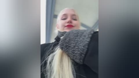 A close-up video of a blonde woman with red lipstick, wearing a black coat and a grey knitted hat, indoors with blurred background.