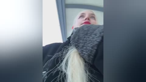 Media: Video of a blonde woman with a black jacket, holding a grey hat with a long, messy, blonde hair tuft sticking out.