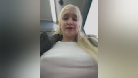 A video of a blonde woman with fair skin, wearing a white top and a dark jacket, sitting in a car with a blurred background.