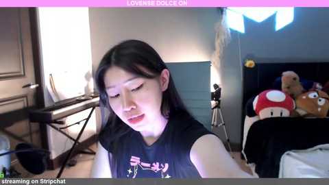 Media: Video of a young Asian woman with long black hair wearing a dark T-shirt, sitting in a dimly lit room with a Super Mario plush toy and a desk in the background.