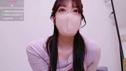 Media: Video of a young Asian woman with long black hair, wearing a white face mask, purple top, and a soft, pale complexion. She's indoors, likely in a bathroom, with a white background.