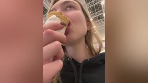 Media: Video of a young woman with light skin and long blonde hair, eating a sandwich with a white crust in a modern indoor setting, blurred background.