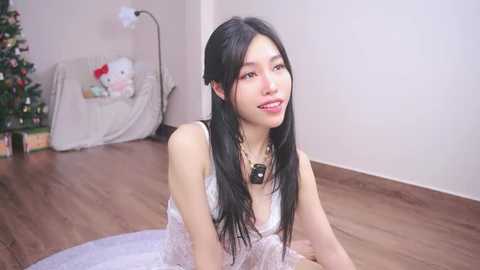 Media: Video of a young Asian woman with long black hair, wearing a white lace dress, sitting on a white rug in a warmly lit room with a Christmas tree and presents in the background.