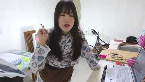 Media: Video of an Asian woman with long black hair, wearing a patterned long-sleeve top and brown skirt, sitting at a cluttered desk with a laptop and phone, smiling while holding a pen.