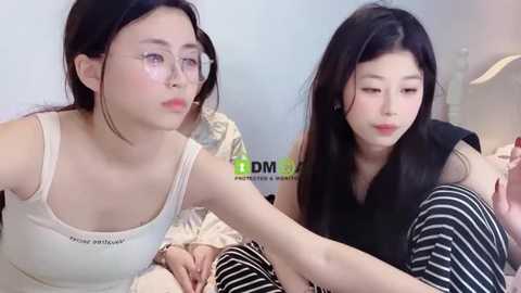 Media: Video of two young East Asian women with fair skin, one in glasses, wearing a white tank top, the other in a black sleeveless top and striped pants, sitting on a bed.