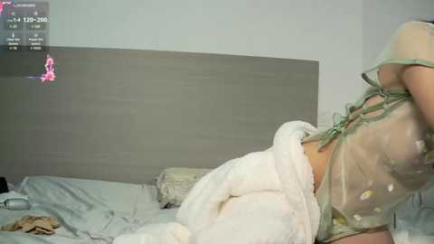 Media: A video of a woman in a sheer, green corset, lying on a bed with a white blanket, in a minimalist bedroom with a grey headboard and a pink notification icon in the top left.