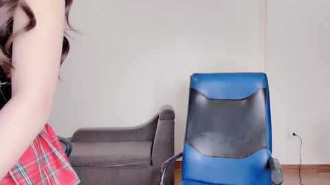 Media: Video of a woman with brown wavy hair in a red plaid shirt, sitting on a gray couch beside a blue gaming chair in a minimalist living room with light walls and wooden floor.