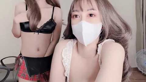 Media: Video of an East Asian woman with light skin, long brown hair, and a white face mask, wearing a black bra and red plaid skirt. Another woman in the background, also masked, wearing a black bra.