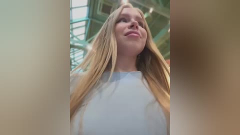 Video of a young woman with long blonde hair, wearing a white top, smiling, taken from a low angle inside a green-tinted, glass-roofed building.