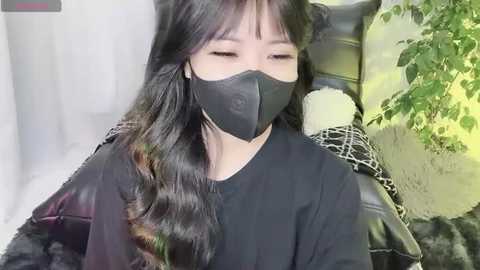 Media: A video of an Asian woman with long, wavy black hair, wearing a black face mask and a black long-sleeved top, seated on a black leather chair, surrounded by greenery.