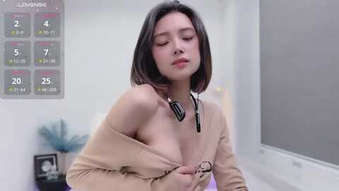 Media: Video of an Asian woman with shoulder-length black hair, wearing a beige off-shoulder sweater, revealing her breasts, in a modern, minimalist room with a gray calendar and plant.