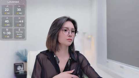 Media: Video of a young woman with shoulder-length dark hair, wearing glasses and a sheer black blouse, sitting indoors with a blurred background, a temperature gauge on the wall.