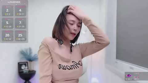 Media: Video of a young Asian woman with shoulder-length black hair, wearing a beige off-shoulder top with \"I love you\" written on it, touching her forehead. Background includes a calendar and minimalistic decor.