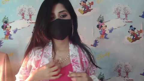 Media: Video of a young South Asian woman with long black hair, wearing a pink floral blouse, black mask, and a gold necklace. Background features Disney characters in a snowy setting.