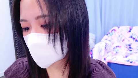 Media: A video of a young Asian woman with straight black hair, wearing a white surgical mask, in a soft, pastel-toned bedroom with a colorful, floral-patterned blanket.